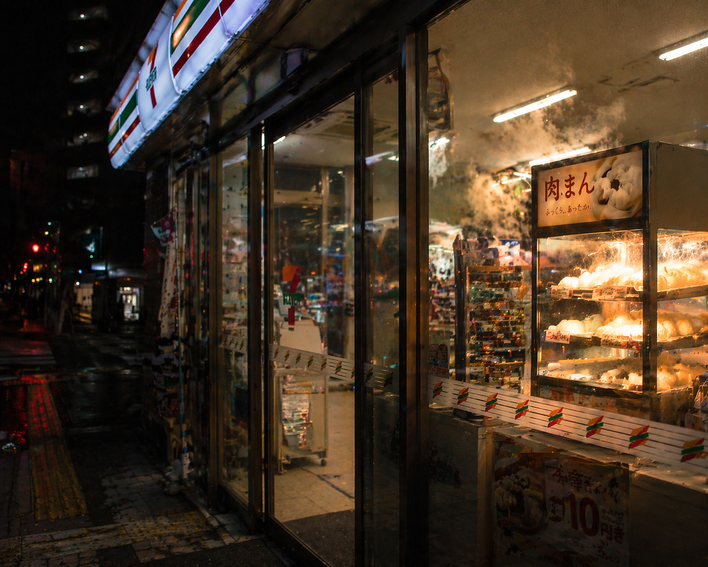 雨上がりの夜道に面したコンビニ。店先の肉まんケースから湯気が上がり、ガラス越しに暖色の光が漏れている。