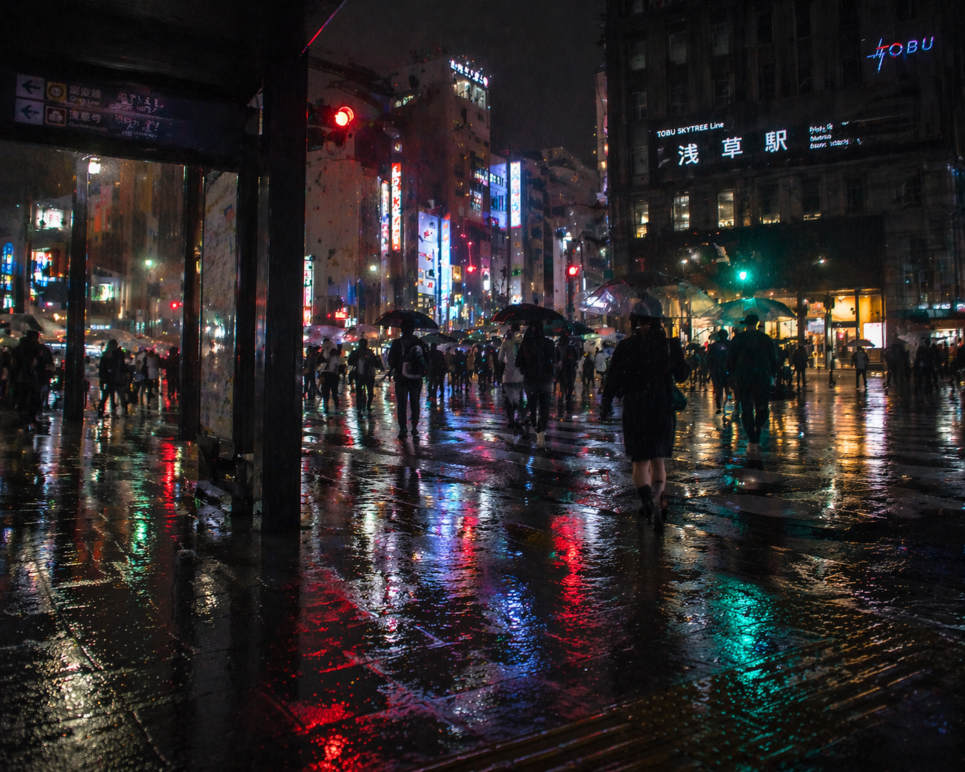 雨の夜の浅草駅前。傘をさした人たちが交差点を渡り、濡れた路面に赤や青の光が反射している。