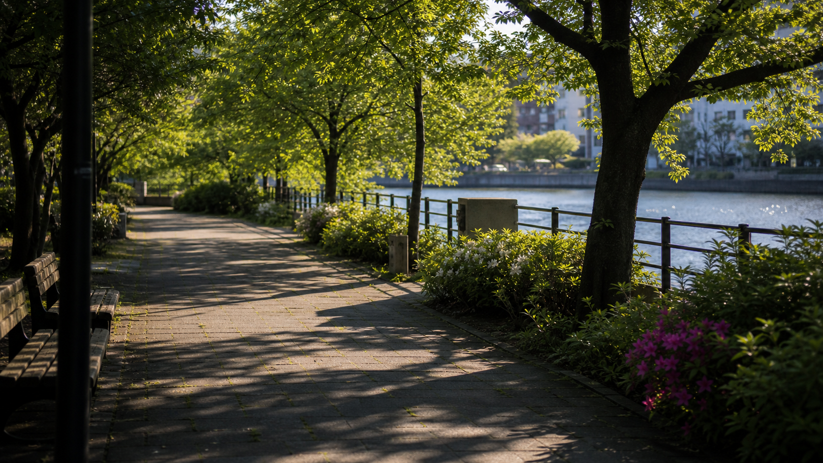 架空の川沿い公園。強い春の光が木陰と遊歩道に落ち、川の気配が葉の間から少し見えている。